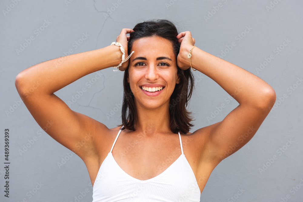 Fototapeta premium smiling young woman with hands on head by gray wall