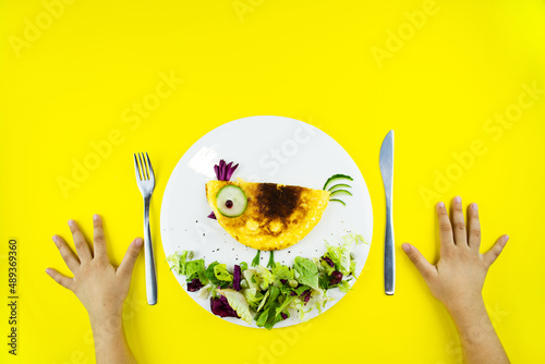Funny chick egg omelette with vegetables and herbs for kids breakfast. Children's hands lie on the table ready to eat.