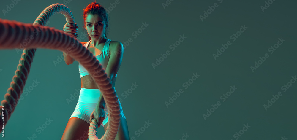 Flyer. Portrait of sportive woman workout, doing exercises with sports equipment isolated on green studio background in neon light. Sport, action, fitness, youth concept. - obrazy, fototapety, plakaty 