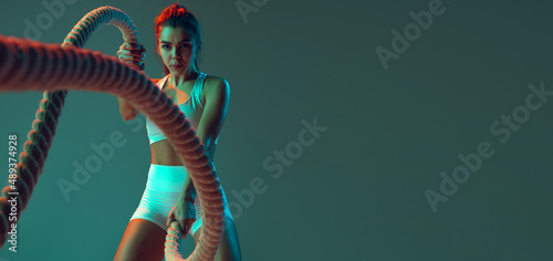 Flyer. Portrait of sportive woman workout, doing exercises with sports equipment isolated on green studio background in neon light. Sport, action, fitness, youth concept.