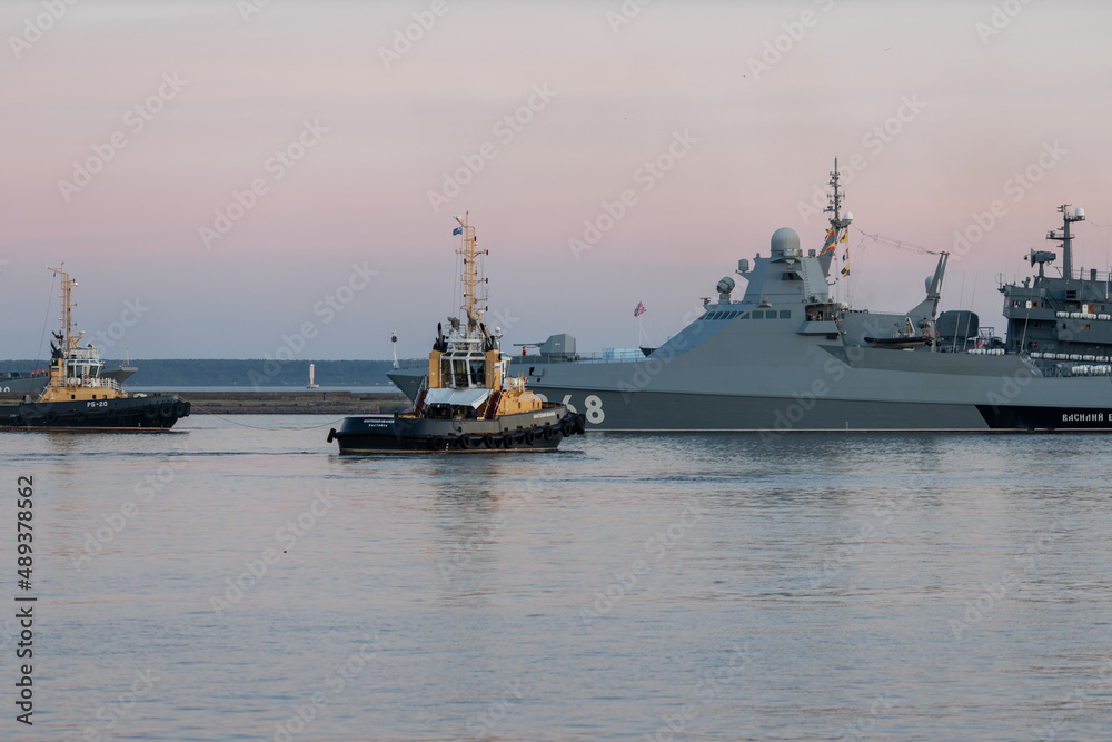 Two naval harbor tugs against the background of warships in the Petrovskaya pier of Kronstadt. Evening sunset. Russia, Kronstadt, July 26, 2020