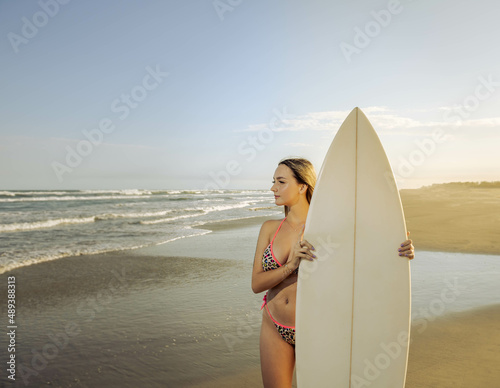 Young beautiful woman standing on the sand looking at the sea with a surfboard in front of her with two in the background. Mention travel, water sports, vacation trips and fun with friends. © studioneeby