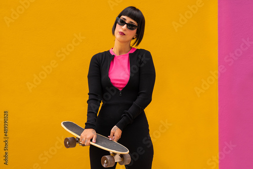 Joven mujer caucásica con tatuajes y un skate sobre un fondo de color