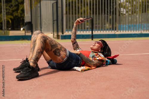 Atractiva mujer con tatuajes en el cuerpo haciéndose unas fotos con la cámara del teléfono en una pista deportiva con un skate