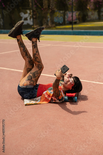 Una mujer caucásica con tatuajes y pelo corto en una pista deportiva con un skate usando la cámara del teléfono
