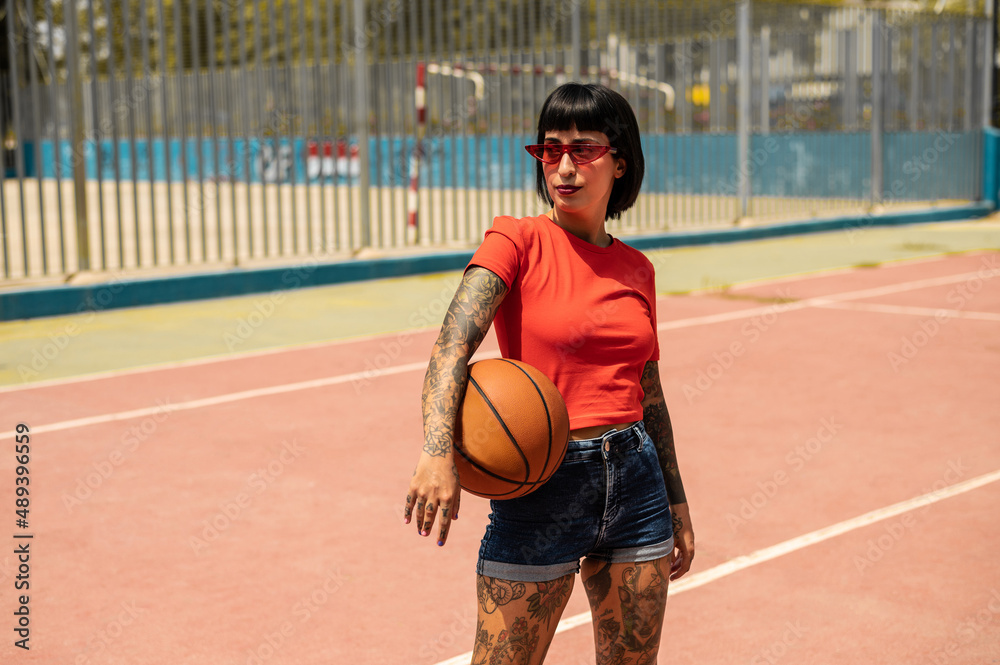 Una mujer caucásica con pelo corto y tatuajes en el cuerpo con un balón de baloncesto	 en una pista deportiva
