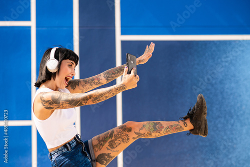 Cheerful woman with tattoos on her arms taking a photo with her phone while listening to music with headphones