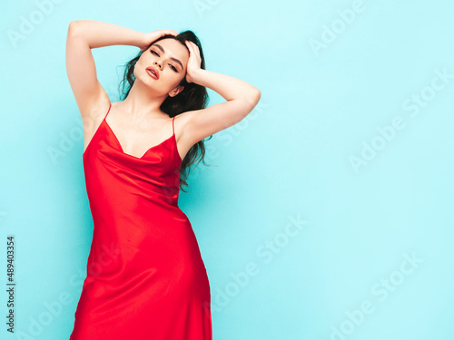 High fashion portrait of young beautiful brunette woman wearing nice trendy red evening dress. Sexy fashion model posing in studio. Fashionable female isolated on blue