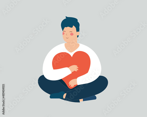 Young man holds, hugs a big heart. Happy adolescent boy sits in lotus pose with closed eyes. Male and cares a red heart with love while sitting. Self acceptance, positive body mental health concept.