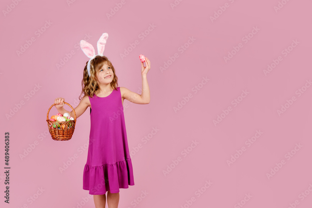 Adorable little girl with blond curly hair in stylish dress and bunny ears headband, smiling and looking away while demonstrating wicker basket and colorful Easter egg, vertical orientation Adorable little girl with blond curly hair in stylish dress and bunny ears headband, smiling and looking away while demonstrating wicker basket and colorful Easter egg, vertical orientation