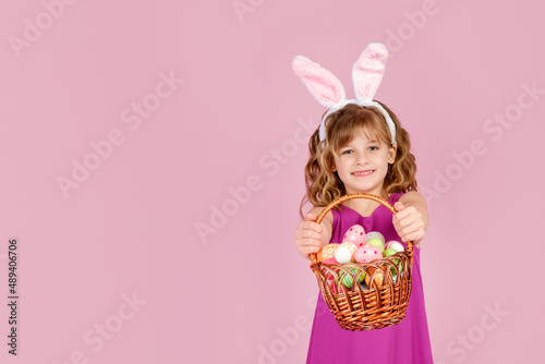 Adorable little girl with blond curly hair in stylish dress and bunny ears, smiling and looking at camera while demonstrating wicker basket full of colorful Easter eggs, copy space