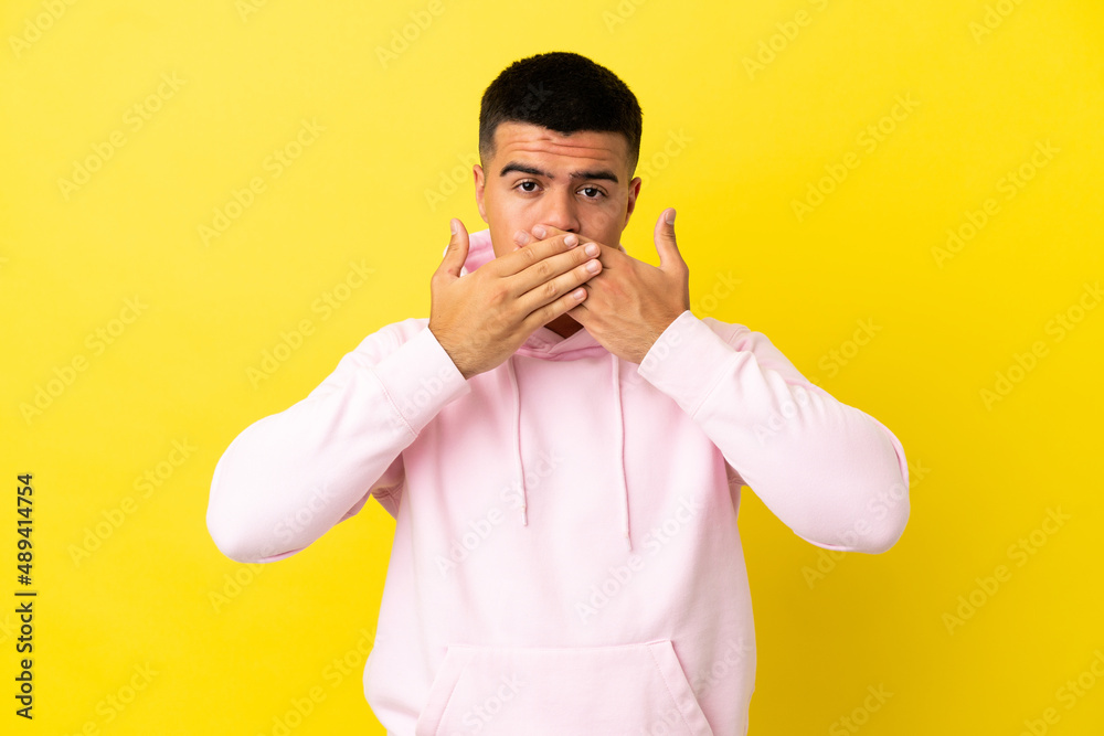 Young handsome man over isolated yellow background covering mouth with hands Young handsome man over isolated yellow background covering mouth with hands