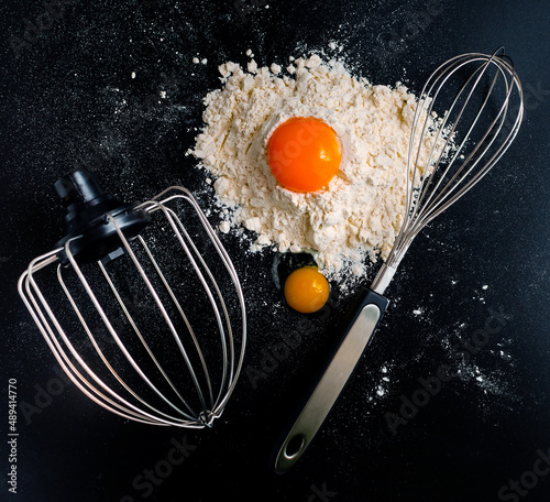 two yolks of chicken and quail eggs with flour and whisks for whipping the dough on a dark background