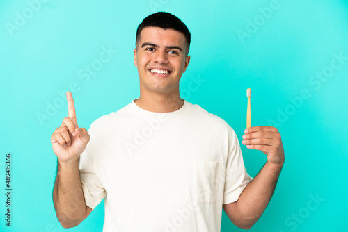Young handsome man brushing teeth over isolated blue background showing and lifting a finger in sign of the best