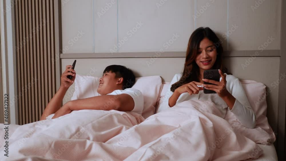 Slow motion shot, Young Couple lying on bed in bedroom, They are enjoy with social media networks addicts surf the internet on mobile phones ignoring each other, family addicted smartphone concept