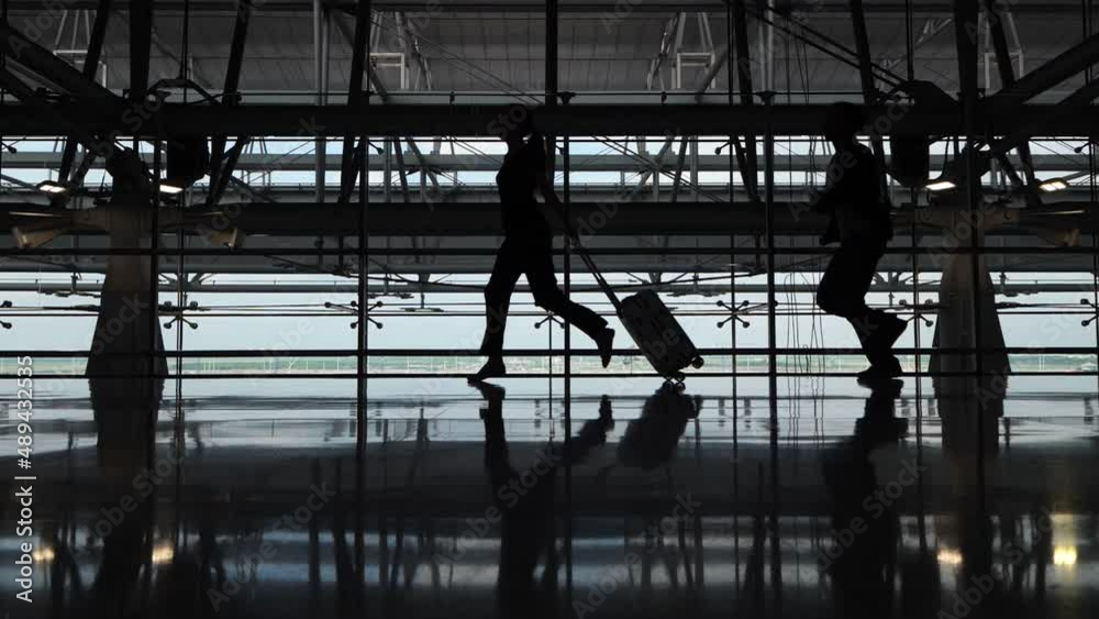 Passengers are late for their flight, a woman runs with a suitcase and a man follows her. Slow motion silhouetted shot, two people running at dim empty hall of terminal Passengers are late for their flight, a woman runs with a suitcase and a man follows her. Slow motion silhouetted shot, two people running at dim empty hall of terminal