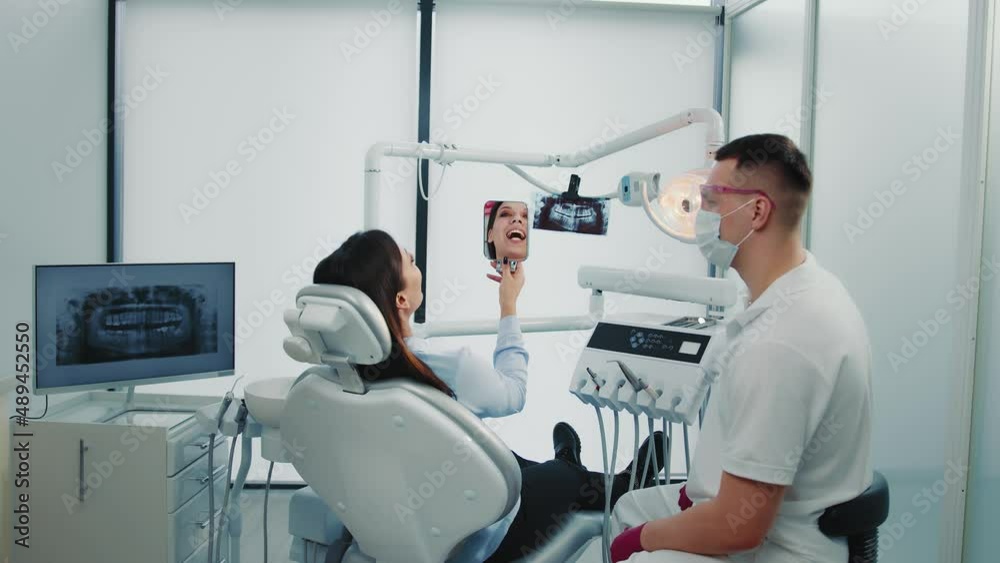 Young woman sitting on chair, looking at mirror, smiling after dentist procedure. Doctor and patient exploring teeth with mirror after treating in dental clinic. Concept of dentistry Young woman sitting on chair, looking at mirror, smiling after dentist procedure. Doctor and patient exploring teeth with mirror after treating in dental clinic. Concept of dentistry