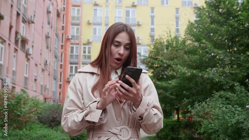 Female in beige coat standing outdoor. She can't believe my eyes using smartphone, smiling and looking surprised, saying wow.