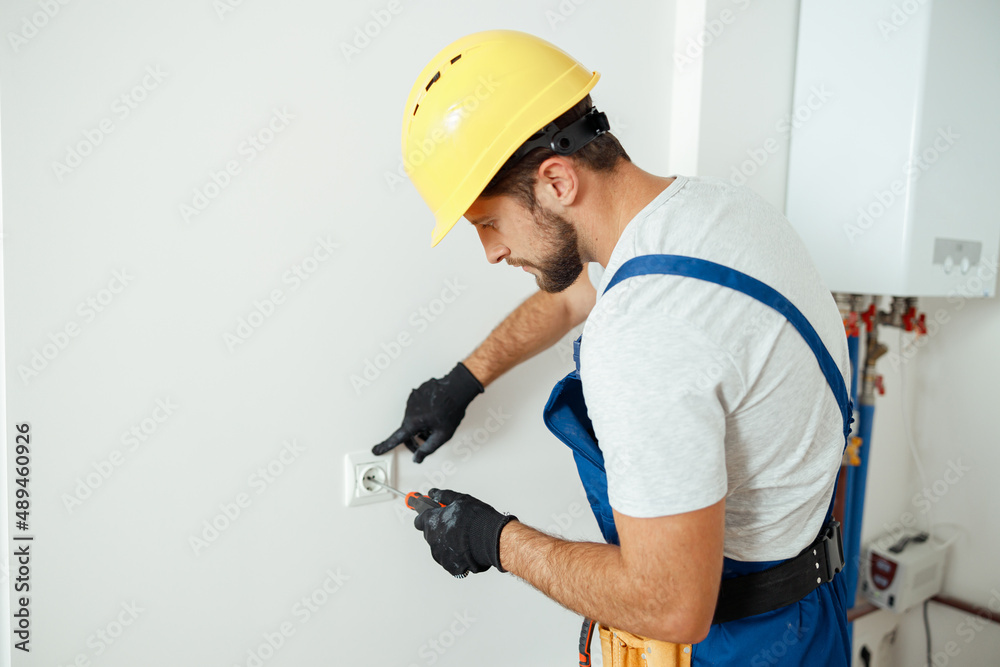 Male worker, professional electrician in hard hat and protective gloves using screwdriver while replacing electrical socket in a room Male worker, professional electrician in hard hat and protective gloves using screwdriver while replacing electrical socket in a room