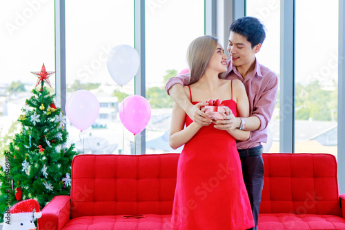 Millennial young Asian lover couple handsome boyfriend giving beautiful girlfriend red present gift box standing smiling cuddling hugging together in living room celebrating valentine day anniversary