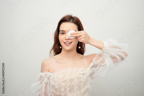 Beauty portrait of a cheerful young topless woman removing face make-up with a cotton pad isolated over gray background