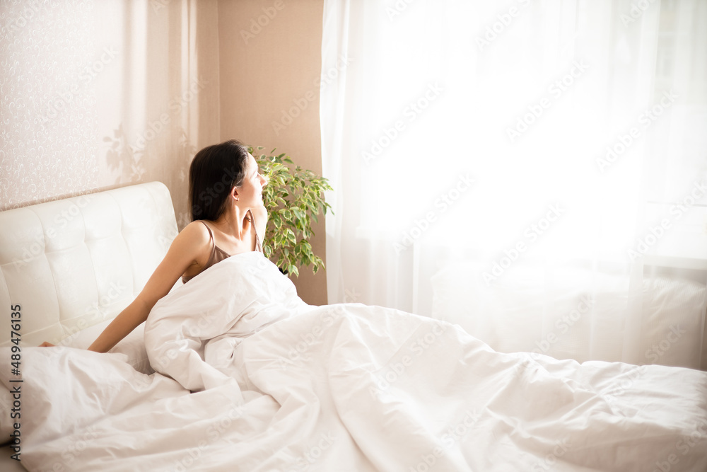 Morning. A young brunette woman wakes up in her bed by the window. Sunlight. Lifestyle