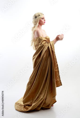  Full length portrait of pretty female model wearing grecian goddess toga gown, posing with elegant gestural movements on a studio background.