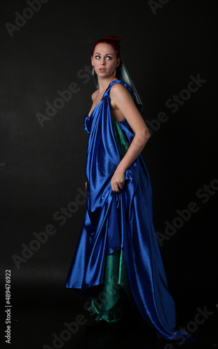  Full length portrait of pretty female model wearing grecian goddess toga gown, posing with elegant gestural movements on a studio background.