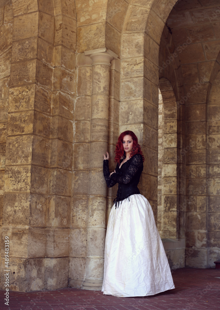 Full length portrait of red-haired woman wearing a  beautiful gothic gown costume, walking around  location with  romantic castle stone architecture background.