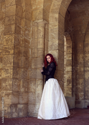 Full length portrait of red-haired woman wearing a beautiful gothic gown costume, walking around location with romantic castle stone architecture background.