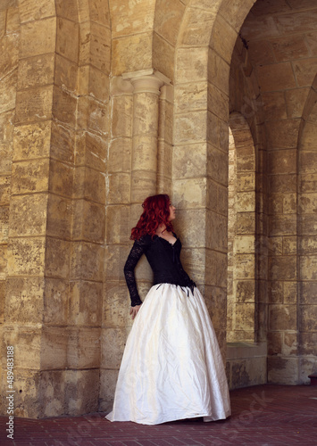 Full length portrait of red-haired woman wearing a  beautiful gothic gown costume, walking around  location with  romantic castle stone architecture background. © faestock