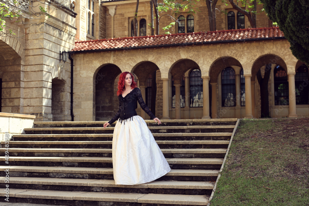 Full length portrait of red-haired woman wearing a  beautiful gothic gown costume, walking around  location with  romantic castle stone architecture background.
