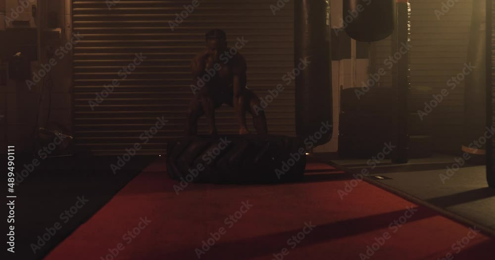 An African American Athlete Exercises, Tyre Flipping In A Crossfit Gym. Shot In An MMA Boxing Gym With Low Key Lighting And A Scattering Of Haze. Captured On Red Digital Cinema Camera An African American Athlete Exercises, Tyre Flipping In A Crossfit Gym. Shot In An MMA Boxing Gym With Low Key Lighting And A Scattering Of Haze. Captured On Red Digital Cinema Camera