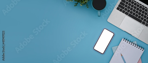 Smart phone, laptop computer, coffee cup and notebook on blue background with copy space.