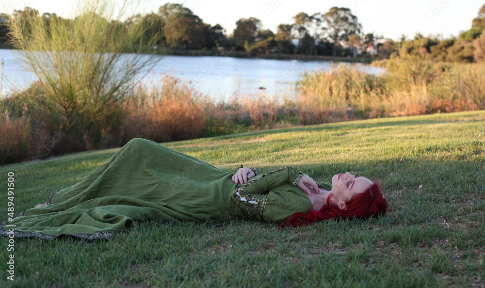 Full length portrait of red haired woman wearing a  beautiful  green medieval fantasy gown. Posing with gestural hands on a enchanted forest background.