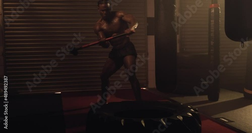 An African American Athlete Exercises, Sledgehammer Tyre Slams In A Crossfit Gym. Shot In An MMA Boxing Gym With Low Key Lighting And A Scattering Of Haze. Captured On Red Digital Cinema Camera 