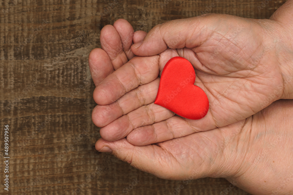 Top view of a flat lay on a wooden table on which an elderly woman folded her plump old hands one on top of the other, putting a red heart in the middle, expressing support, love and understanding. Top view of a flat lay on a wooden table on which an elderly woman folded her plump old hands one on top of the other, putting a red heart in the middle, expressing support, love and understanding.