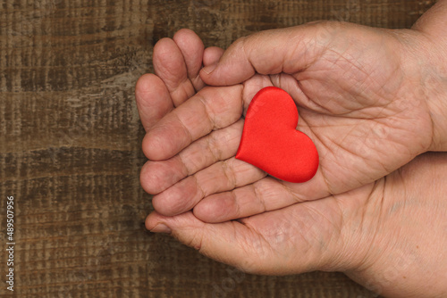 Top view of a flat lay on a wooden table on which an elderly woman folded her plump old hands one on top of the other  putting a red heart in the middle  expressing support  love and understanding.