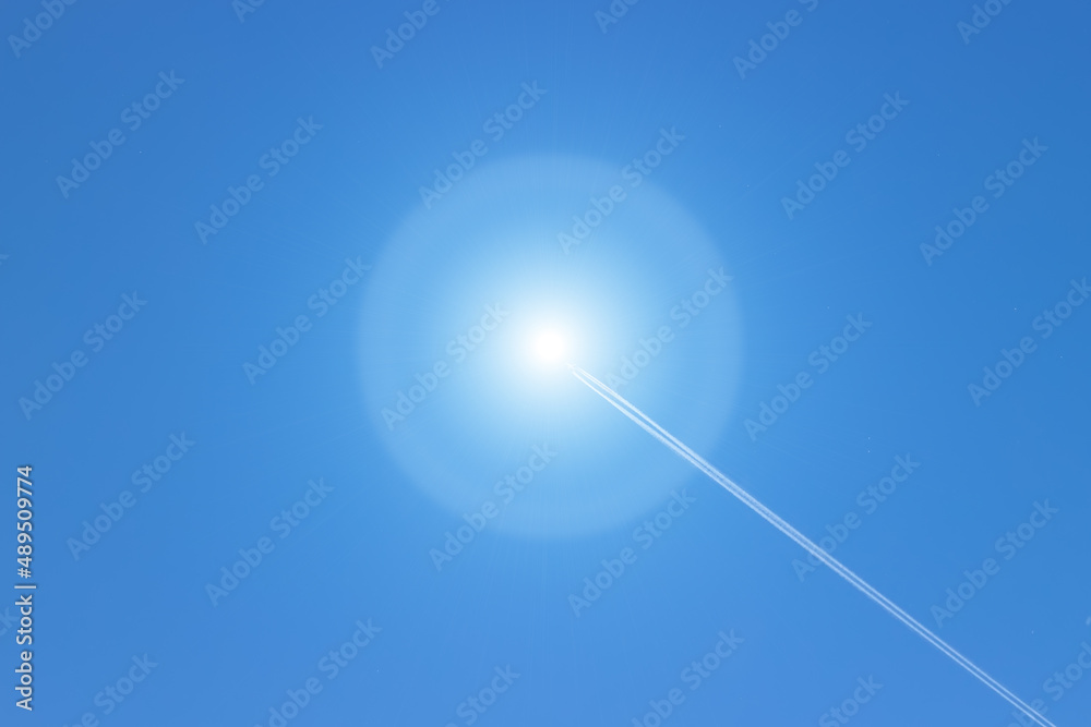 White fluffy condensing trail from the plane ends in the sun against a bright blue sky. Solar flare in the sky with vapor trail from aircraft. White fluffy condensing trail from the plane ends in the sun against a bright blue sky. Solar flare in the sky with vapor trail from aircraft.