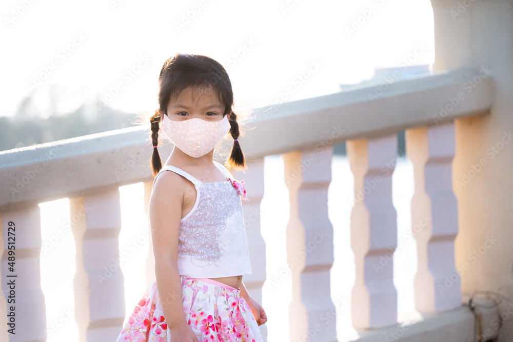 Portrait adorable kid girl wearing cloth face mask travel public park in evening. Summer or spring time. Child stand and watch nature at foot of bridge across pond. Beautiful children aged 5 years old