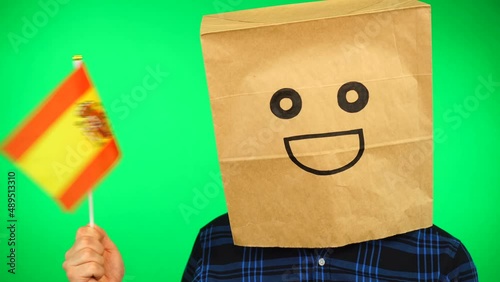 Portrait of man with paper bag on head waving Spanish flag with smiling face against green background.