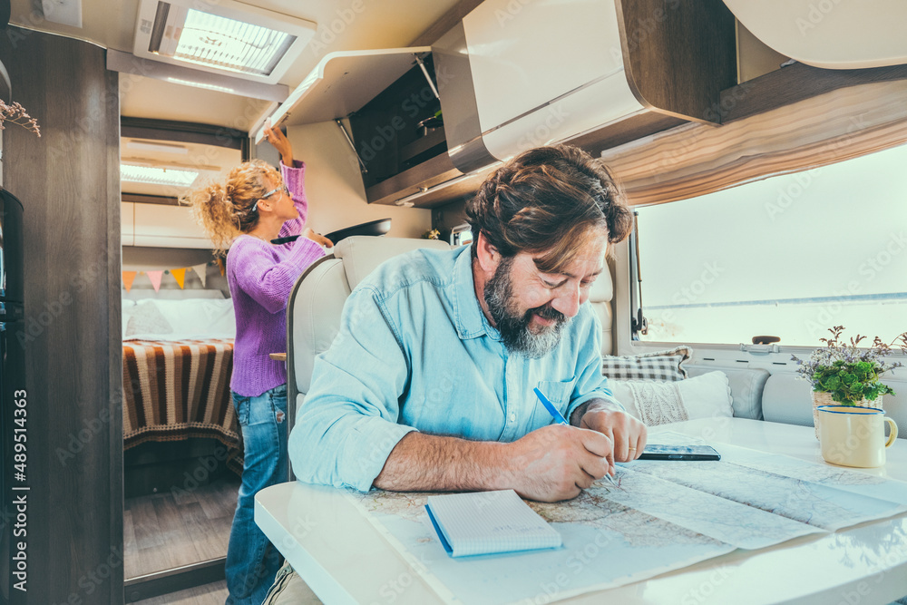 Bearded man planning next destination travel holiday vacation sitting at the table inside his camper van. Woman in background cooking and managing storage. Holiday life vacation Bearded man planning next destination travel holiday vacation sitting at the table inside his camper van. Woman in background cooking and managing storage. Holiday life vacation
