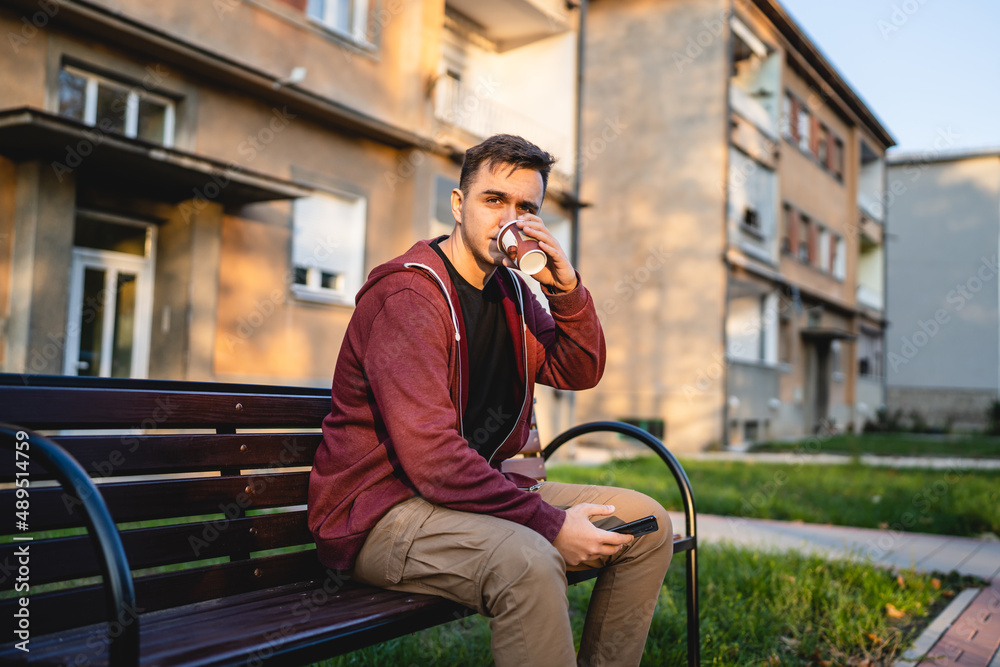 Obraz premium One young adult caucasian man sitting outdoor on the bench in autumn day holding mobile phone and a cup of coffee looking to the side alone waiting making a phone call or sms in front of building