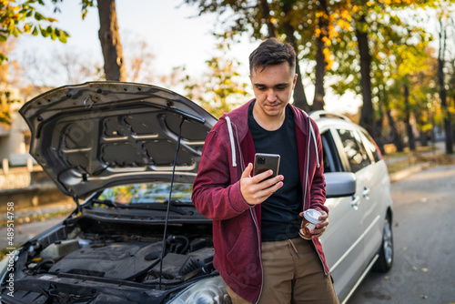 One young adult caucasian man standing by his vehicle with open hood and broken failed engine holding a phone calling towing service for help on the road Roadside assistance concept in autumn day