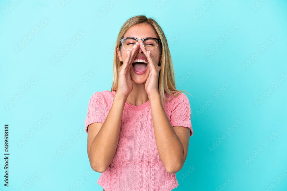 Blonde Uruguayan girl isolated on blue background shouting and announcing something Blonde Uruguayan girl isolated on blue background shouting and announcing something