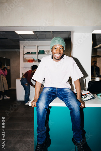 Portrait of happy young man sitting on dining table in kitchen at college dorm photo