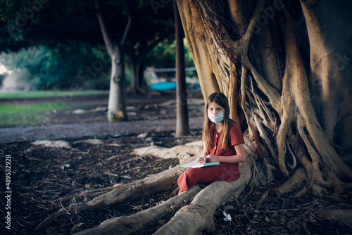 A cute european baby girl in a dress and a mask sits near a big tree in the park. A child among the big roots of a tree writes poetry  a girl makes notes
