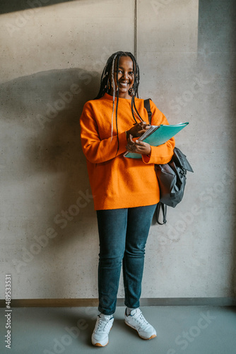 Full length portrait of smiling young woman holding books and backpack standing against gray wall