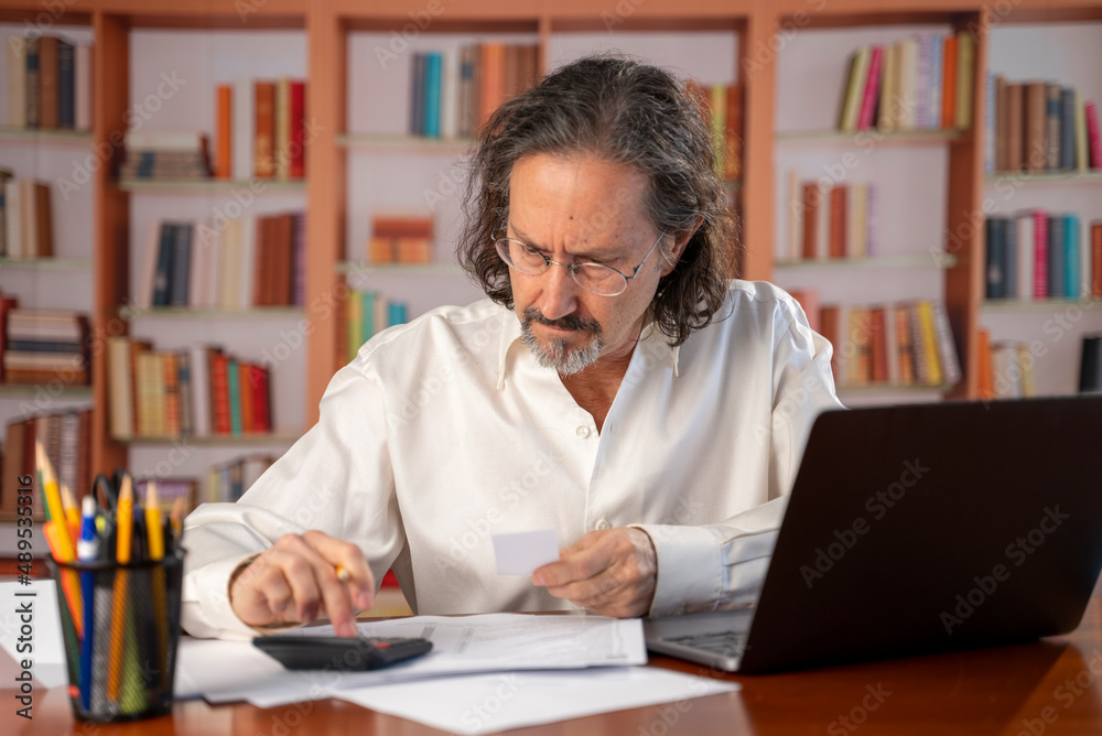 Serious man wearing glasses with beard checking bills, using calculator, holding receipt, sitting at table with laptop, accountant calculating company expenses for financial report, planning budget Serious man wearing glasses with beard checking bills, using calculator, holding receipt, sitting at table with laptop, accountant calculating company expenses for financial report, planning budget