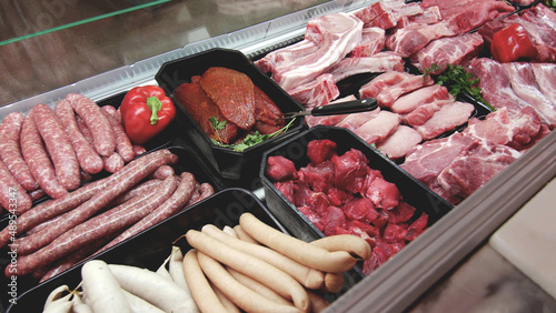 meat department display shelves with selection of typical fresh raw meats slices and variety of sausage products inside market or supermarket refrigerated background.